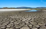 Cracked, dry and exposed lake bottom in foreground of bed of Lower Klamath Lake, California with small patch of remaining water in background against backdrop of blue sky and distant barren hills.