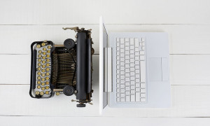 Overhead shot of and old fashioned typewriter back-to-back with a modern laptop computer on w white wood desk.