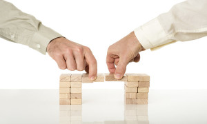 Conceptual image of business merger and cooperation - two male hands joining effort to build a bridge of wooden pegs on a white desk with reflection over white background.