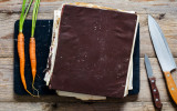 Vintage cook book. Old cook book with healthy recipes on wooden cutting board. Two young fresh carrots and two kitchen knifes on sides. Top view, copy space