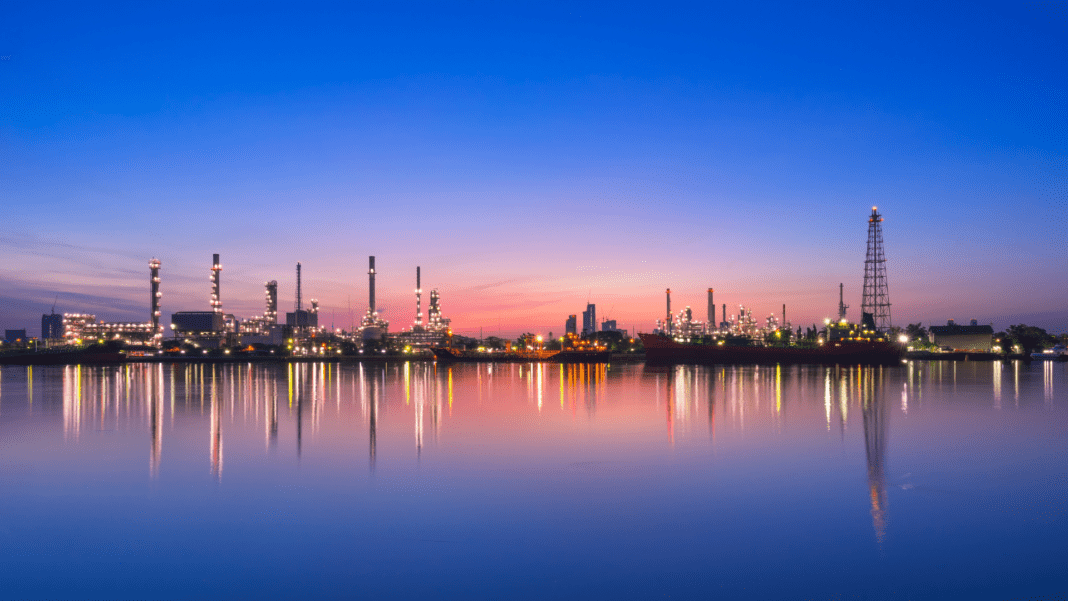 image2 An oil refinery silhouetted against a vibrant sunset, with its reflection shimmering on the calm water below.
