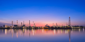 An oil refinery silhouetted against a vibrant sunset, with its reflection shimmering on the calm water below.