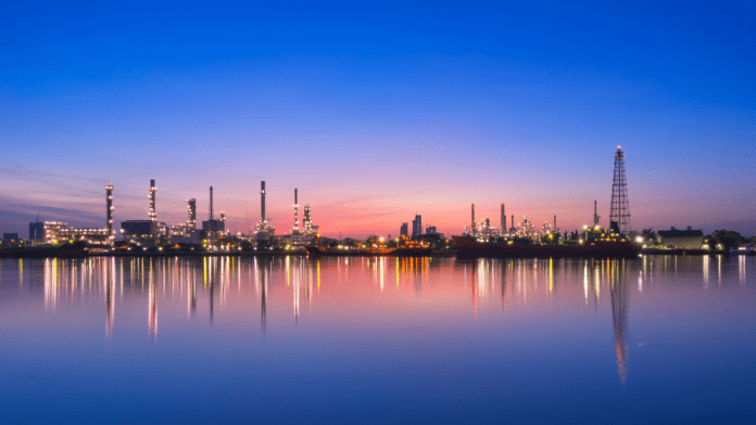 An oil refinery silhouetted against a vibrant sunset, with its reflection shimmering on the calm water below.