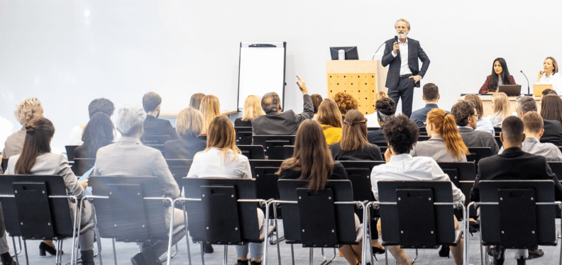 A speaker presenting to an audience in a conference room with attendees raising their hand to ask a question