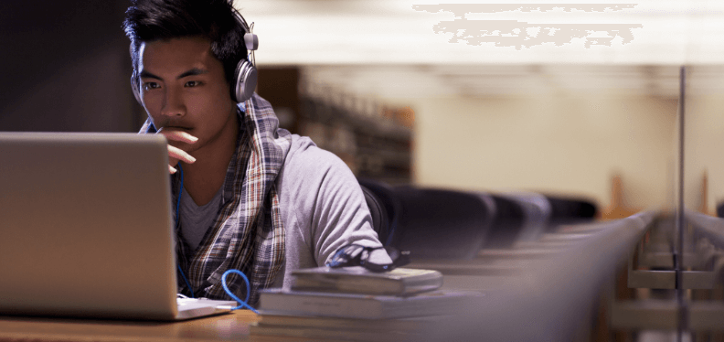 A focused young man wearing headphones while using a laptop in a library setting