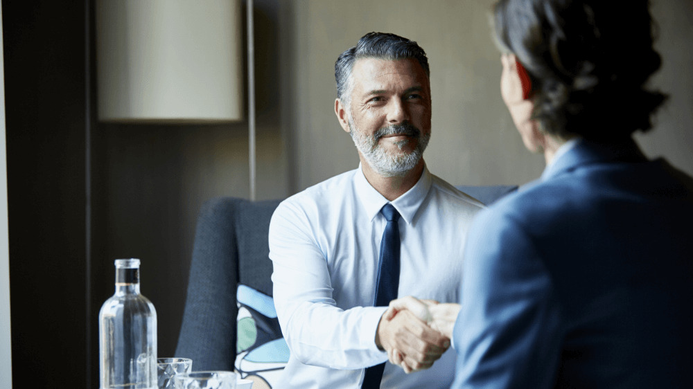 Two professionals in a meeting, shaking hands across a table, implying a deal or agreement